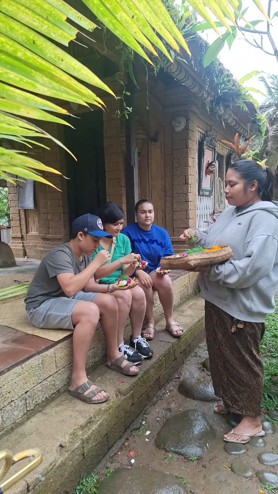 Guests tasting Balinese snacks offered by a local woman in a village compound, 2026